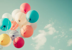 Colorful party balloons attached to white ribbon float against a turquoise sky with wispy white clouds